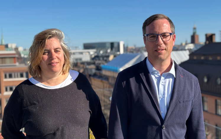 Två personer står på ett tak med stadslandskap och blå himmel i bakgrunden.
