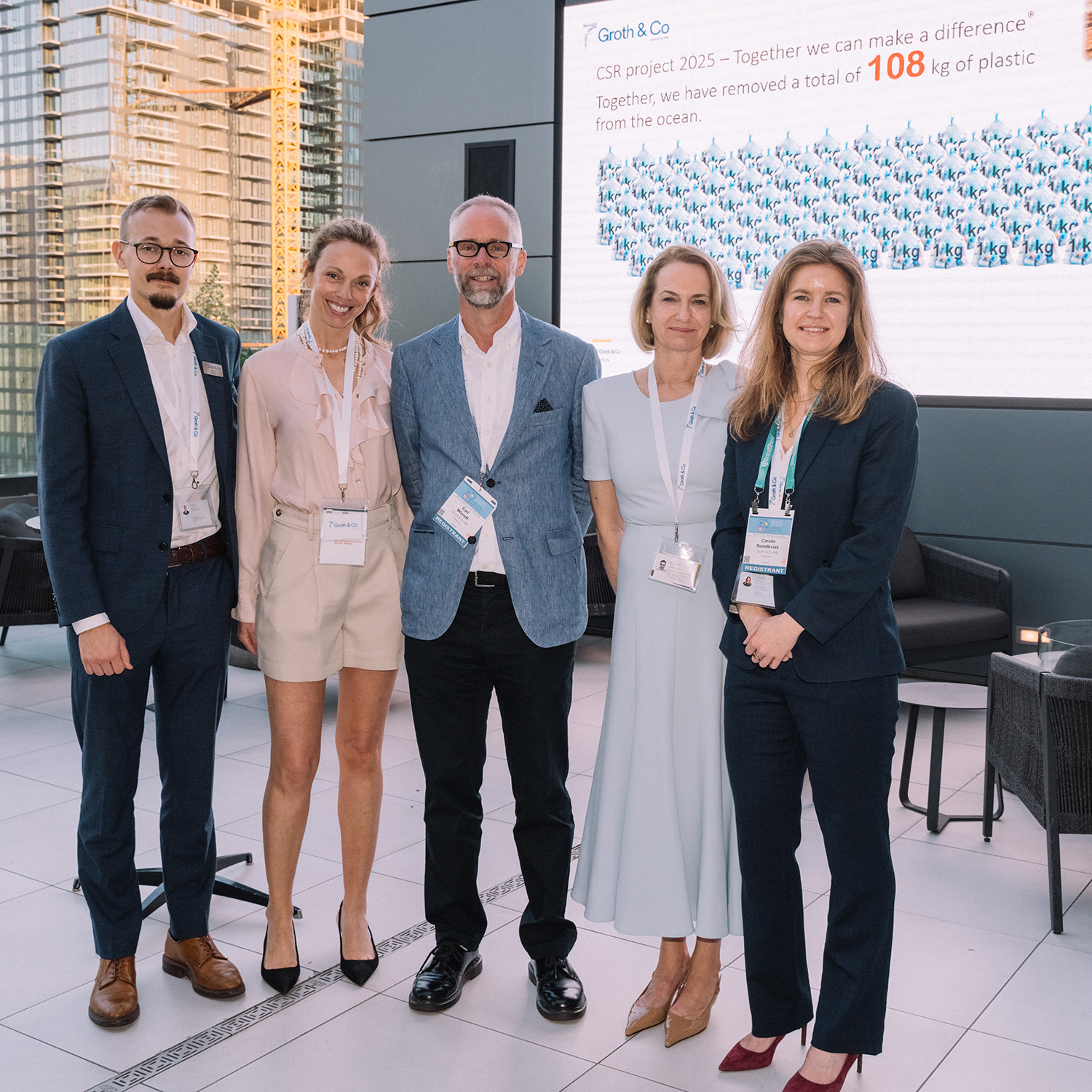 Fem personer poserar tillsammans vid ett evenemang med stor skärm i bakgrunden.